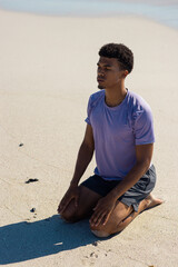 Obraz premium At beach, young biracial man sitting on sand, looking away thoughtfully