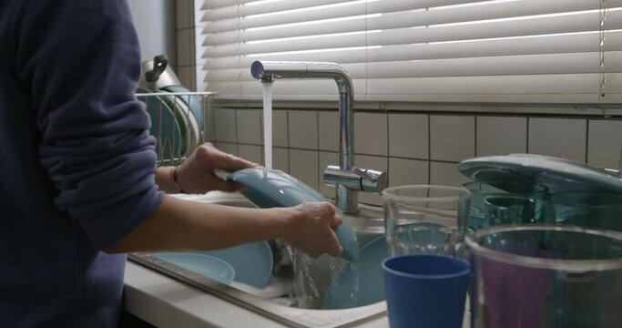 A woman is standing at a kitchen sink, washing dishes under running water from a faucet. Sunlight is streaming in from a window with blinds.