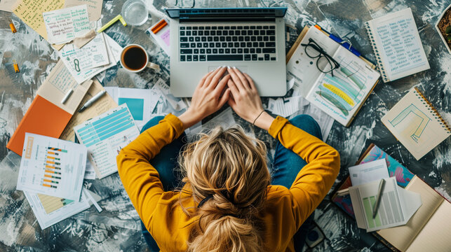 Top view of a hectic woman working on an urgent project for work on her laptop with papers with sheets and graphs and data and a cup of coffee and her glasses are around her