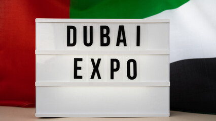 Lightbox with text DUBAI EXPO on background of waving UAE flag made from silk. United Arab Emirates flag with concept of tourism and traveling. Inviting greeting card, advertisement. Dubai welcoming