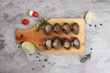 Arrangement Of Raw Blood Clams On Wooden Cutting Board With Lemon And Herbs Over Grey Background