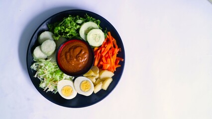 Gado-gado served on a plate, isolated white, Indonesian salad containing boiled vegetables and potatoes, boiled eggs, fried tofu tempeh, lontong and melinjo chips, served with peanut-cashew sauce. 