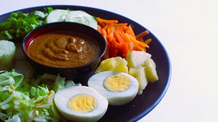 Gado-gado served on a plate, isolated white, Indonesian salad containing boiled vegetables and potatoes, boiled eggs, fried tofu tempeh, lontong and melinjo chips, served with peanut-cashew sauce. 