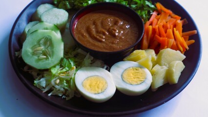 Gado-gado served on a plate, isolated white, Indonesian salad containing boiled vegetables and potatoes, boiled eggs, fried tofu tempeh, lontong and melinjo chips, served with peanut-cashew sauce. 