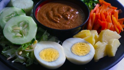 Gado-gado served on a plate, isolated white, Indonesian salad containing boiled vegetables and potatoes, boiled eggs, fried tofu tempeh, lontong and melinjo chips, served with peanut-cashew sauce. 