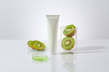 An empty label cosmetic tube in white color displayed in center of photo, next to a petri dish of chopped kiwi, a beaker and petri dish with fresh kiwi in back. Mock up for designing, vacant space