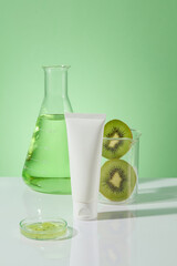 Over the green background, an unbranded tube in white color is displayed with a conical flask of liquid, a beaker of kiwi slices and a petri dish. Mockup of skin care cosmetic tube of beauty facial