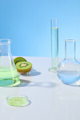 Baby blue background against some laboratory instrument filled by transparent liquid placed on white counter, decorated by green color of kiwi. Photo of front view, vacant space for advertising