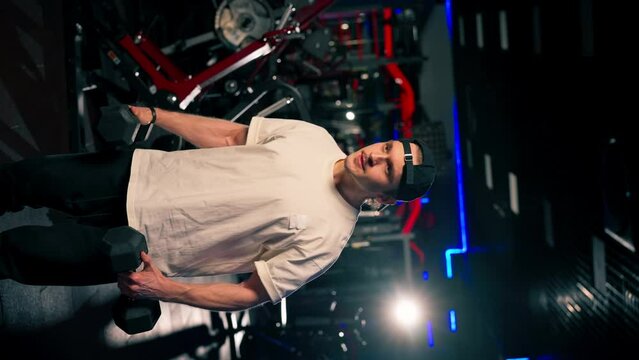 vertical video in the gym young handsome guy in a T-shirt and a cap does exercises with dumbbells and shakes his arms