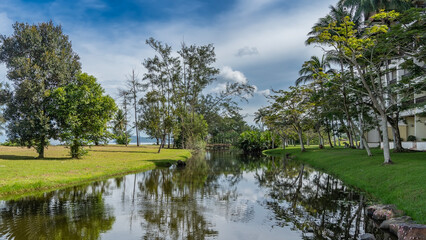 Fototapeta premium A calm river in a tropical park. Sprawling trees grow on the banks. Green grass on the lawns. A fragment of the hotel building in the distance. Clouds in the blue sky. Reflection. Malaysia. Borneo