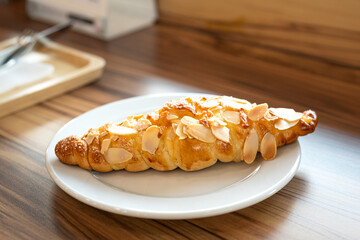 A piece of baked croissant with cheese and almond topping in a white plate and placed on wooden...