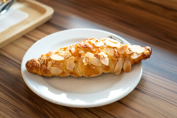 A piece of baked croissant with cheese and almond topping in a white plate and placed on wooden...