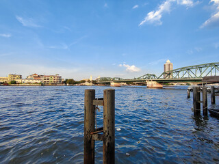  Phra Phuttha Yot Fa Bridge over the Chao Phraya River. The bridge is painted in green and features a truss design. In the foreground, there are wooden posts sticking out of the water, showing signs o