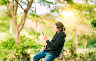 Young man sitting on a swing using cell phone in nature. Smiling guy sitting on a swing with smart phone