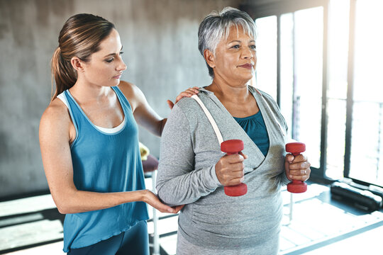 Senior woman, dumbbells and help with coach in gym for physio, stretching arms and rehabilitation exercise. Old female patient, instructor support and workout for arthritis with balance and weights