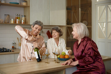 Excited retired ladies in pajamas uncorking bottle of wine at home kitchen