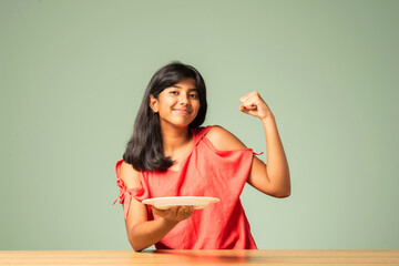 Asian Indian adorable girl child holding empty ceramic plate with happy expressions, sitting on...