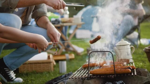 Hands of unrecognizable father and son flipping sausages and burgers while grilling in nature during camping