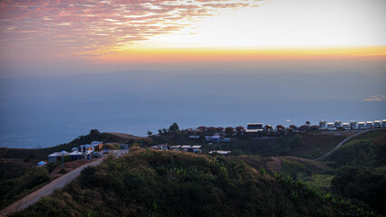 picturesque landscape at dusk. A winding road snakes through a hilly terrain adorned with lush greenery. On the right, there is a cluster of buildings, possibly a small village or resort, perched on t