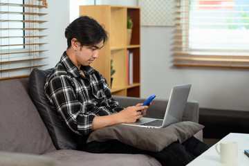 Focused young man using laptop and smartphone on couch in living room
