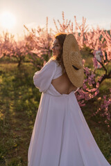 Woman blooming peach orchard. Against the backdrop of a picturesque peach orchard, a woman in a long white dress and hat enjoys a peaceful walk in the park, surrounded by the beauty of nature.