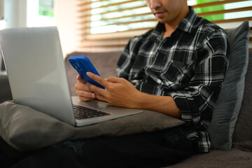 Cropped shot male freelancer sitting on sofa with laptop and using mobile phone