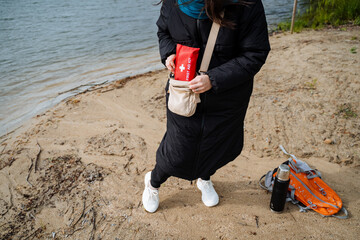 Woman by lakeside beach in warm coat and white sneakers, holding red first aid kit backpack....