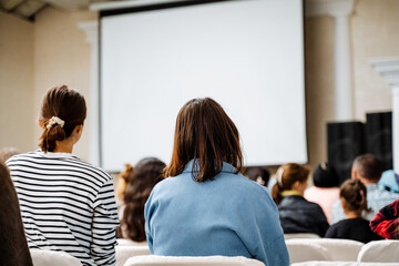 Audience watching a presentation in a seminar room with a white screen background. People at a...