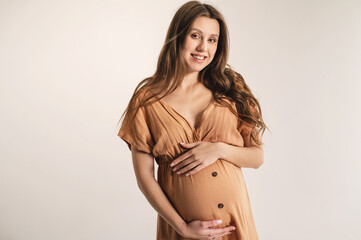 A young pregnant woman in an peach fuzz summer dress on a light plain background. Portrait of a smiling positive pregnant woman with long hair