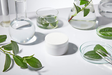 Laboratory equipment with green tea ingredient flat lay on white table top, an unlabeled cosmetic jar featured in center, sample for adding brand packaging. Mock up for design, high angle shot