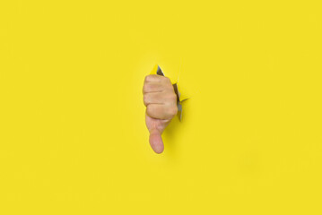 Male hand with thumb down in sign of disapproval, appearing out of the hole in a torn yellow paper...