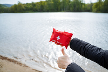 Person holds bright red first aid kit, giving thumbs up by serene lake. Ready for outdoor...