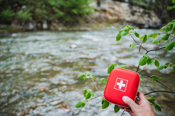 Hand holding a red and white cross first aid kit by River Outdoor Safety and Preparedness in lush...