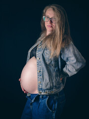 A pregnant woman wearing glasses stands sideways against a black background. Her unbuttoned denim...