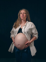 The pregnant woman stands against a dark background, creating a striking contrast. She is dressed...