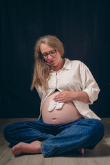 A pregnant in casual attire, sitting cross-legged on floor, holds a pair of baby socks against...