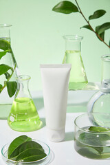 Close-up photo of a white unlabeled cosmetic tube placed on a green and white background with some laboratory equipments and green tea decorated around. Mock up for promotion, space for text