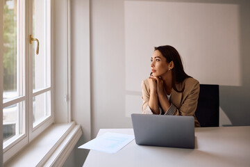 Pensive businesswoman looking through window while working on laptop in office.