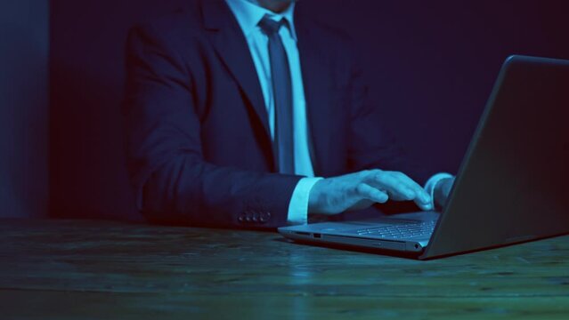 A man in a business suit with a tie works at a table with a laptop in the evening. Dark blue scheme. The hand pushing an imaginary button or icon.