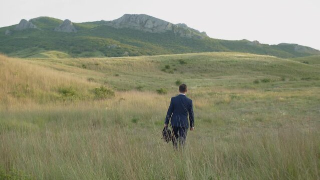 Man in suit with suitcase in nature walk in tall grass to the mountains during sunset. The idea of revaluation of true values.