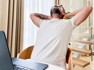 Tired man for overwork stretching his back in front of an open laptop in hotel room. Traveler and...