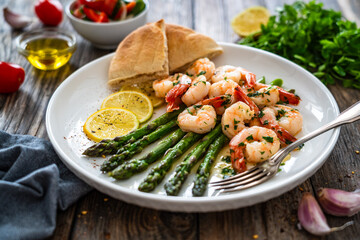 Fried shrimps with garlic and parsley in olive oil and cooked green asparagus on wooden table
