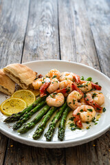 Fried shrimps with garlic and parsley in olive oil and cooked green asparagus on wooden table
