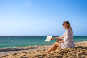 Mid-adult beautiful woman sitting on sunny beach by the sea reading book
