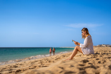 Mid-adult beautiful woman sitting on sunny beach by the sea reading book
