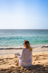 Mid-adult beautiful woman sitting on sunny beach by the sea reading book
