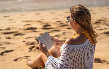 Mid-adult beautiful woman sitting on sunny beach by the sea reading book
