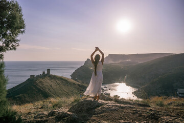 woman stands on a rocky hill overlooking a body of water. She is wearing a white dress and she is...