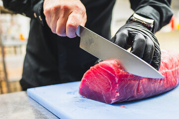 A person in black attire with black gloves slices a large piece of tuna on a blue cutting board...