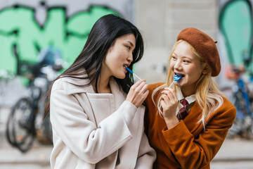 Lesbian couple looking at each other while eating lollipops during a date outdoors.